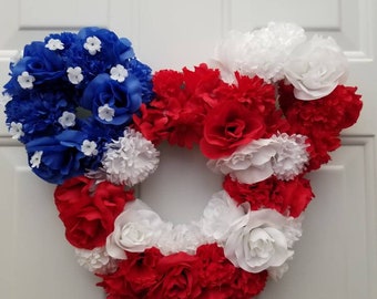May include: A red, white, and blue floral wreath in the shape of a Mickey Mouse head. The wreath is made of artificial flowers and is perfect for a patriotic display.