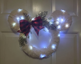 May include: A white faux fur Mickey Mouse-shaped Christmas wreath with a red and black plaid bow and green sprigs. The wreath is decorated with white LED lights.