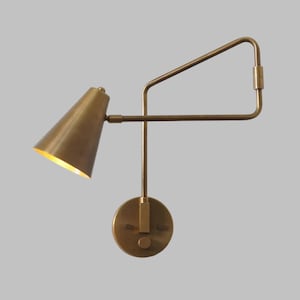 May include: A brass wall-mounted light fixture with a cone-shaped shade and an adjustable arm. The light is on and casting a warm glow.
