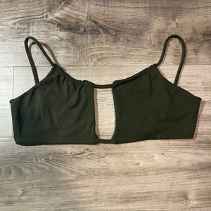 May include: Dark green crop top with thin spaghetti straps and a keyhole cutout. The top is made of a smooth, solid fabric and is designed to be worn as a top. The garment is laid flat on a wooden surface.