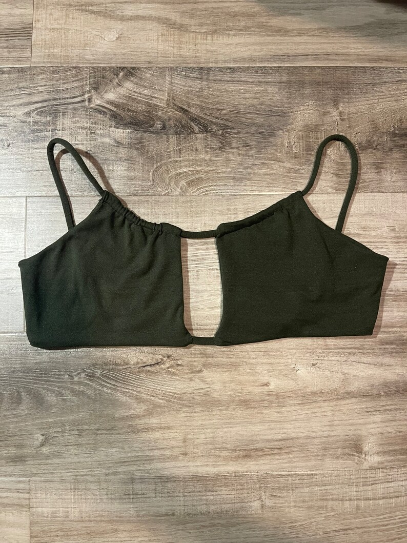 May include: Dark green crop top with thin spaghetti straps and a keyhole cutout. The top is made of a smooth, solid fabric and is designed to be worn as a top. The garment is laid flat on a wooden surface.