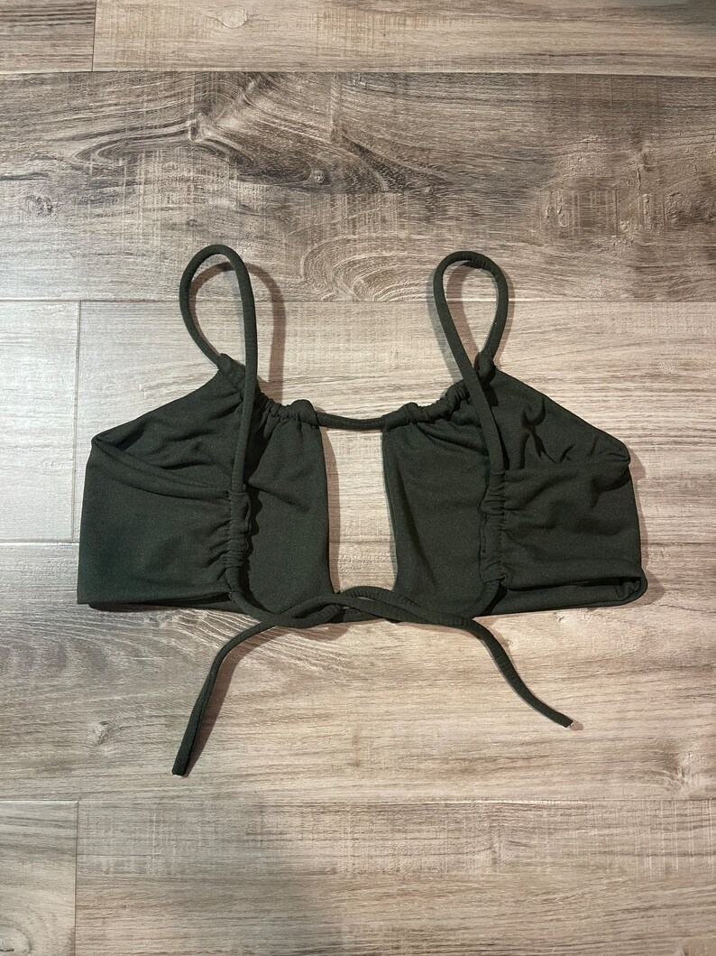 May include: Dark green crop top with thin straps and a keyhole design. The top features gathered fabric details on the sides and ties at the bottom. The top is made of a soft, stretchy material.