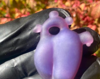 May include: A translucent, lavender-colored pendant with a unique, abstract design. The pendant has a rounded body with a central hole and small, wing-like protrusions. The object is held against a background of colorful foliage.