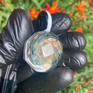 May include: A faceted glass pendant with a black and white top. The pendant features swirls of blue, yellow, and white within a clear glass body. The pendant is held in a gloved hand against a backdrop of green foliage and orange flowers.