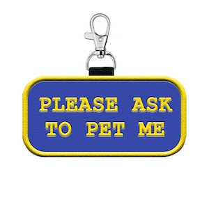 May include: A blue rectangular tag with yellow text that reads "PLEASE ASK TO PET ME." The tag has a yellow border and a silver clasp attached to a black strap. The tag is designed to be attached to a pet's collar.