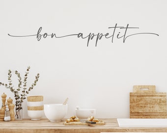 May include: A kitchen wall decal with the text "bon appétit" in a cursive font. The decal is in dark gray and is placed above a kitchen counter with a wooden countertop and a vase of greenery.