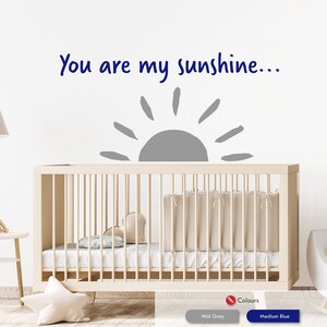 Peut inclure: Un autocollant mural de cr&egrave;che avec un soleil gris et le texte "You are my sunshine..." en bleu. L'autocollant est plac&eacute; au-dessus d'un lit b&eacute;b&eacute; blanc avec un matelas blanc et une couverture beige.