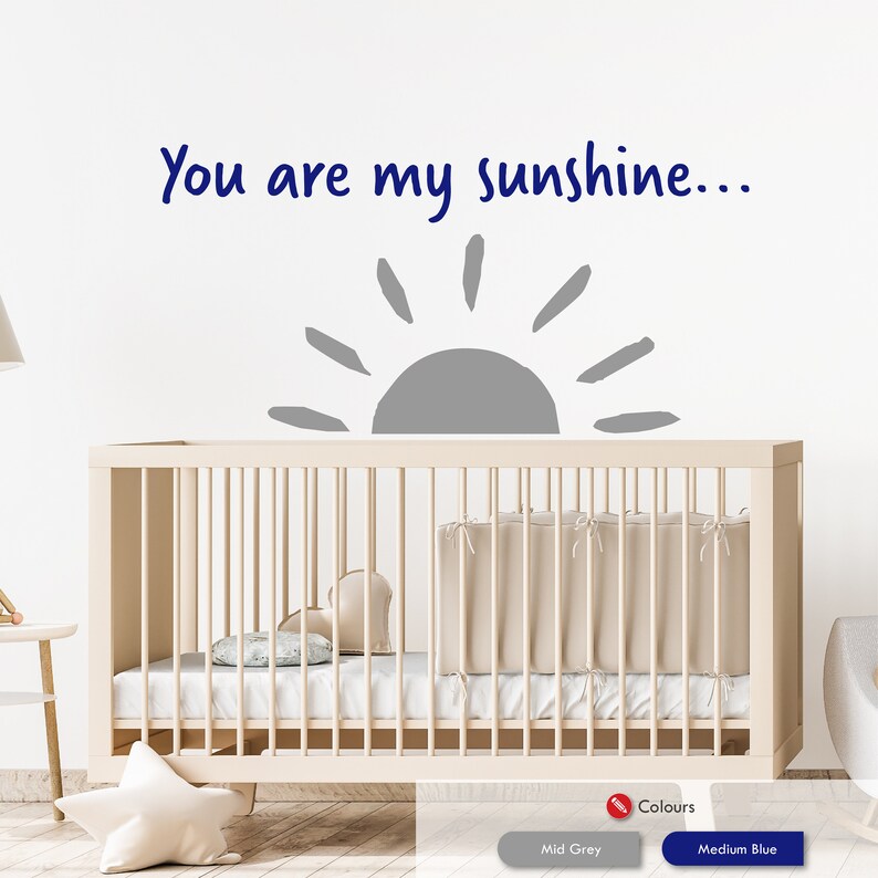 Peut inclure: Un autocollant mural de cr&egrave;che avec un soleil gris et le texte "You are my sunshine..." en bleu. L'autocollant est plac&eacute; au-dessus d'un lit b&eacute;b&eacute; blanc avec un matelas blanc et une couverture beige.