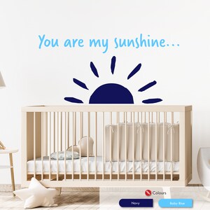 Peut inclure: Un autocollant mural de cr&egrave;che avec un soleil bleu et le texte "You are my sunshine..." en bleu clair. L'autocollant est plac&eacute; au-dessus d'un lit b&eacute;b&eacute; blanc avec un matelas blanc et une couverture blanche.