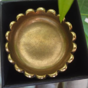 Puede incluir: Un plato pequeño, redondo, de color dorado con un borde festoneado. El plato está hecho de metal y tiene una superficie lisa y brillante.
