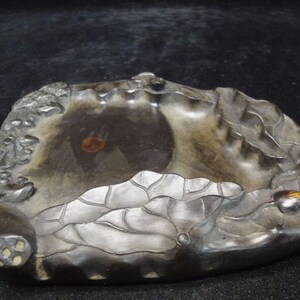 May include: A dark gray, carved stone ink well with a lotus flower design. The ink well has a central basin and is decorated with detailed relief carvings of lotus leaves and flowers. The overall shape is organic and irregular.