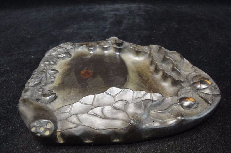 May include: A dark gray, carved stone ink well with a lotus flower design. The ink well has a central basin and is decorated with detailed relief carvings of lotus leaves and flowers. The overall shape is organic and irregular.