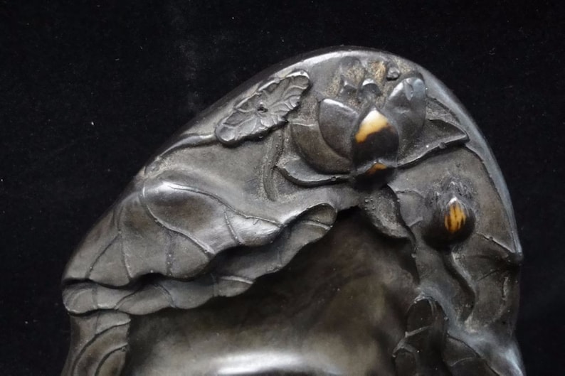 May include: Close-up of a dark, possibly metal, decorative object with a lotus flower and leaf design. The lotus flower has a light-colored center, and the leaves are intricately detailed.