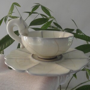 May include: Cream-colored ceramic teacup and saucer set. The teacup has a unique handle and sits on a leaf-shaped saucer. The set has a delicate, crackled pattern. The background features green leaves.