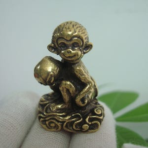 Può includere: Una piccola statuetta di scimmia in ottone dorato seduta su una nuvola. La scimmia tiene un oggetto rotondo nella sua mano sinistra.