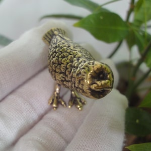 May include: A small, gold-colored metal bird figurine with detailed textured feathers. The bird has a round head with a small beak and large, round eyes.
