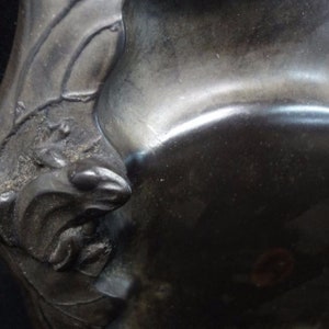 May include: Close-up of a dark, possibly bronze, sculpture. The image focuses on a detailed section, revealing a textured surface with a relief of a bat or similar creature. The sculpture has a smooth, reflective area.