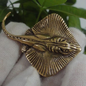 Puede incluir: Una figurita de metal de color dorado que representa una raya con detalles precisos. La raya está representada con sus alas extendidas y su cola enroscada.