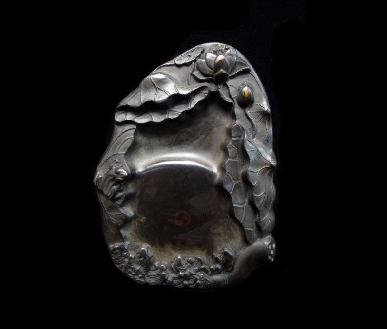 May include: A silver-toned, antique inkwell with a lotus flower and leaf design. The inkwell has a shallow, oval-shaped basin and is decorated with detailed relief carvings of aquatic plants and flowers.