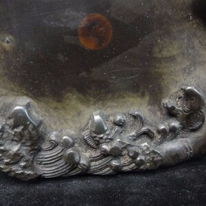 May include: A dark, metallic-toned decorative dish with a detailed relief design. The relief features stylized waves and aquatic creatures. The dish has a smooth, reflective surface with a circular amber-colored spot.