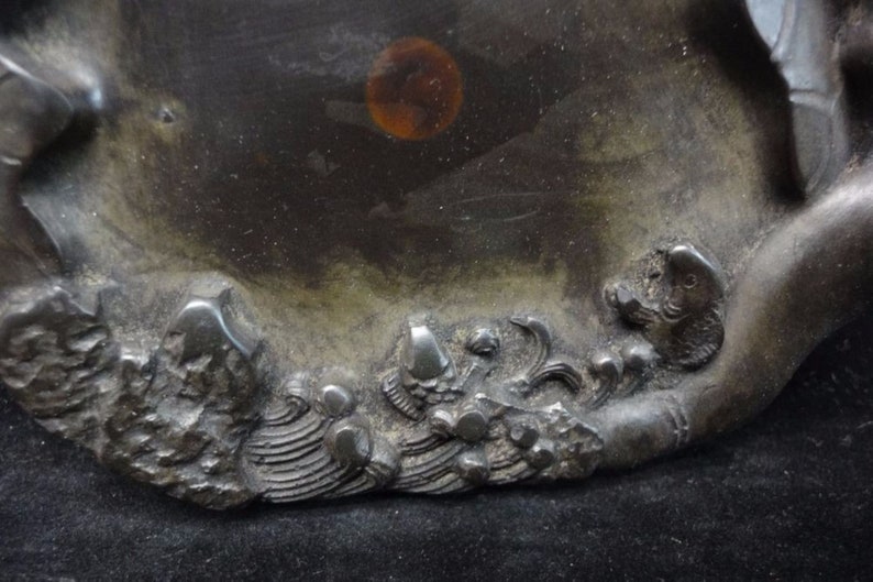 May include: A dark, metallic-toned decorative dish with a detailed relief design. The relief features stylized waves and aquatic creatures. The dish has a smooth, reflective surface with a circular amber-colored spot.
