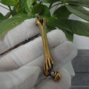 May include: A gold-colored bone-shaped pendant with a small, clear stone at the end. The pendant is hanging from a white cloth.