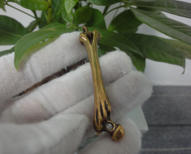May include: A gold-colored bone-shaped pendant with a small, clear stone at the end. The pendant is hanging from a white cloth.