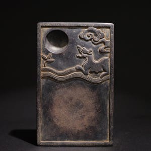 Puede incluir: Piedra de tinta rectangular gris oscuro con relieve tallado. Presenta una luna llena, nubes estilizadas y una criatura mítica sobre líneas onduladas. Hay un pozo circular para la tinta. La piedra tiene un borde enmarcado.