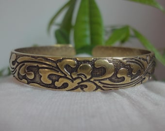 Puede incluir: Un brazalete de metal dorado con un intrincado diseño floral. El brazalete es liso y tiene un acabado pulido.