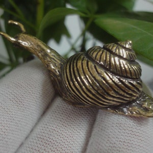 May include: A gold-coloured snail figurine with a detailed shell and a textured body. The snail is positioned on a white surface.