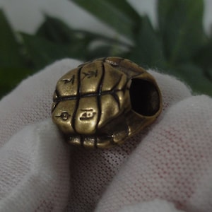 Könnte beinhalten: Nahaufnahme einer kleinen Messingperle im antiken Stil. Die Perle hat die Form eines Schildkrötenpanzers mit geätzten Linien und Symbolen. Die Perle hat ein Loch, das hindurchführt, und wird in einem weißen Tuch gehalten.