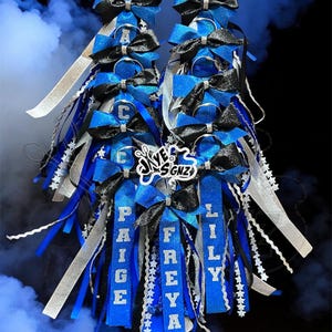 Può includere: Una decorazione homecoming mum con strati di nastri blu e neri glitterati, accenti argentati e finiture a forma di stella. I nastri riportano i nomi PAIGE, FREYA e LILY in lettere bianche, con un logo centrale che recita JAYE SGNZ.