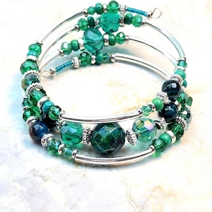 May include: A spiral bracelet with three silver bands, adorned with various shades of green beads. The beads include faceted, round, and small seed beads, creating a layered and textured look. The bracelet is designed to wrap around the wrist.