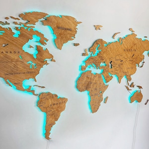 Puede incluir: Un mapa del mundo de madera con un brillo azul claro alrededor de los continentes. El mapa est&aacute; montado en una pared blanca y tiene un cable enchufado. El mapa incluye los nombres de pa&iacute;ses y continentes, incluyendo "Groenlandia", "Canad&aacute;", "Estados Unidos de Am&eacute;rica", "Rusia", "Brasil" y "Australia".