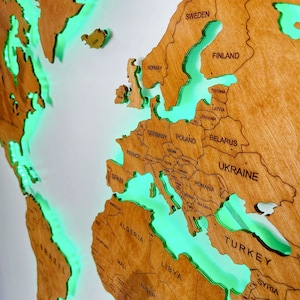 Puede incluir: Un mapa del mundo de madera con un brillo verde alrededor de los continentes. El mapa est&aacute; montado en una pared blanca y muestra los pa&iacute;ses de Europa, &Aacute;frica y partes de Asia y Am&eacute;rica del Sur.