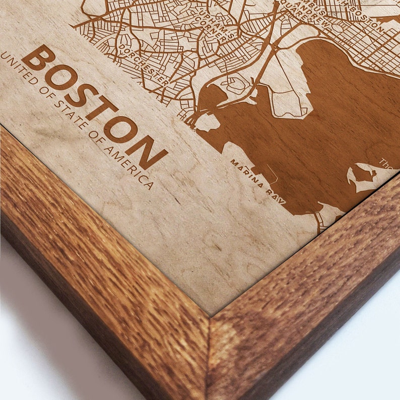 May include: A framed wooden map of Boston, Massachusetts, with the city's streets and waterways engraved in brown on a beige background. The text "BOSTON" and "UNITED OF STATE OF AMERICA" is printed in brown at the top left corner of the map. The text "MARINA BAY" is printed in brown at the bottom right corner of the map.