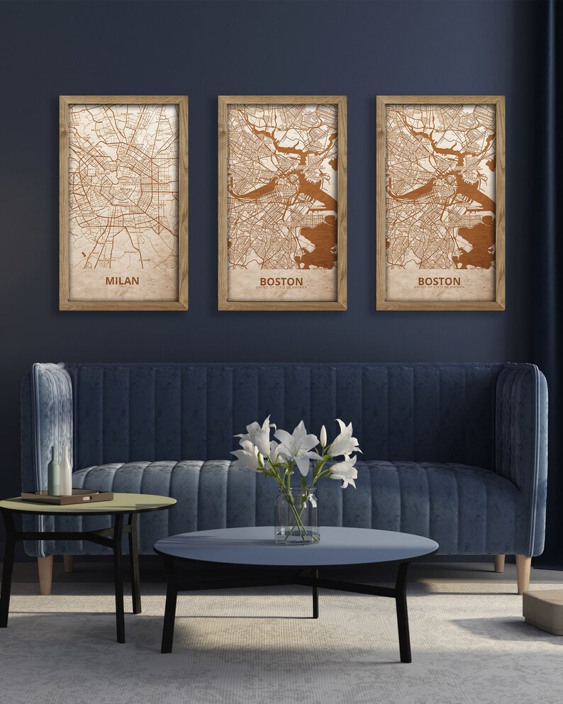 May include: Three framed maps of Milan and Boston, with a brown background and copper-colored lines, hanging on a dark blue wall above a blue velvet sofa and two coffee tables.