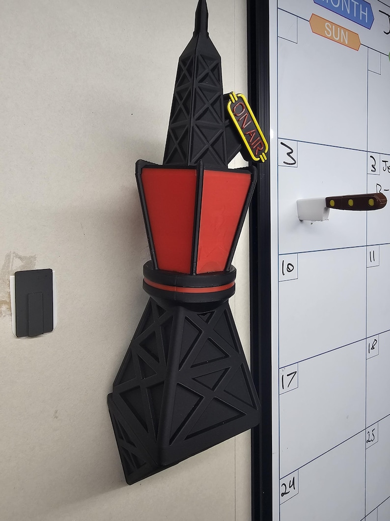 Puede incluir: Un im&aacute;n negro y rojo con forma de torre de radio con un letrero amarillo de "ON AIR". El im&aacute;n est&aacute; adherido a una pared blanca. La torre tiene un dise&ntilde;o geom&eacute;trico y una secci&oacute;n central roja.