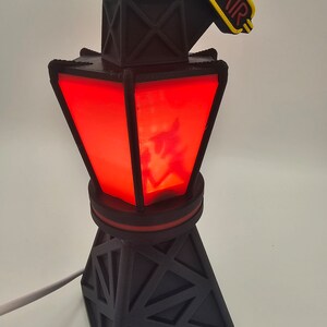 Puede incluir: L&aacute;mpara de torre de radio negra impresa en 3D con un centro rojo iluminado. La l&aacute;mpara tiene un letrero "ON AIR" en amarillo y rojo. La l&aacute;mpara est&aacute; enchufada con un cable blanco.