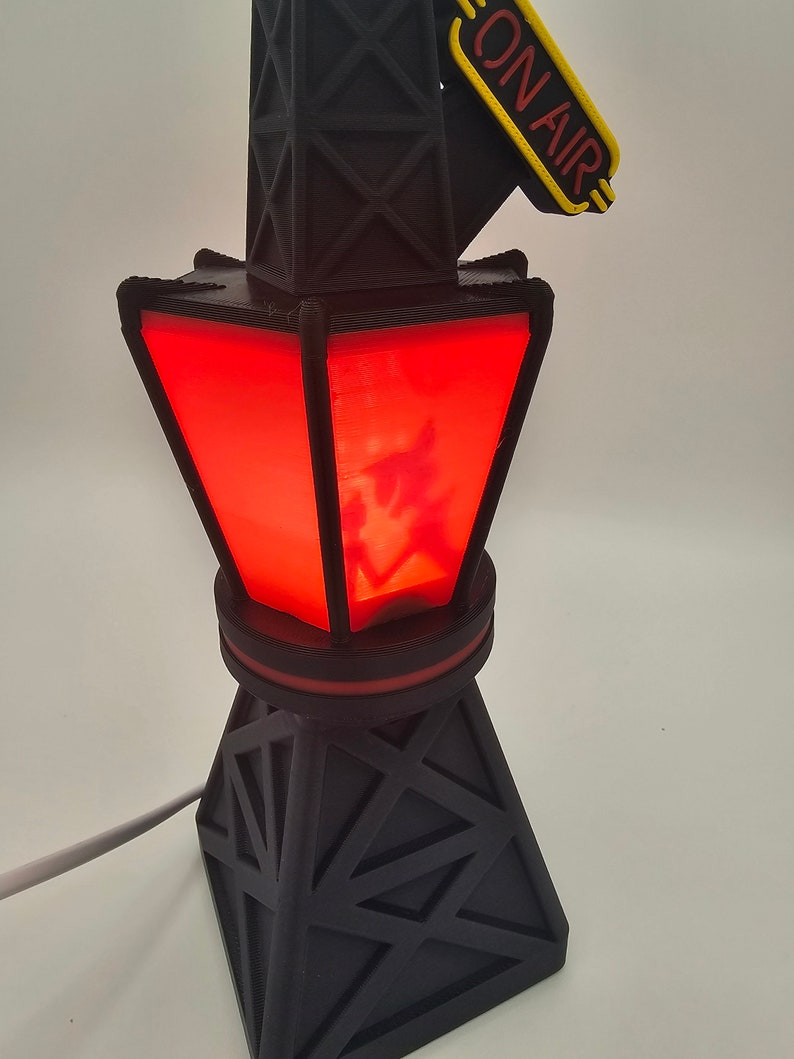Puede incluir: L&aacute;mpara de torre de radio negra impresa en 3D con un centro rojo iluminado. La l&aacute;mpara tiene un letrero "ON AIR" en amarillo y rojo. La l&aacute;mpara est&aacute; enchufada con un cable blanco.