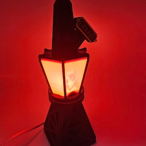 Puede incluir: Una l&aacute;mpara negra con forma de torre con un resplandor rojo que emana del interior. La l&aacute;mpara tiene un letrero de "ON AIR". La base tiene forma de pir&aacute;mide geom&eacute;trica. La l&aacute;mpara est&aacute; enchufada a una pared roja.