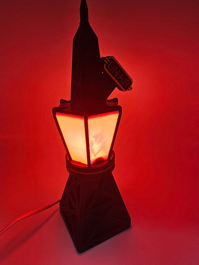 Puede incluir: Una l&aacute;mpara negra con forma de torre con un resplandor rojo que emana del interior. La l&aacute;mpara tiene un letrero de "ON AIR". La base tiene forma de pir&aacute;mide geom&eacute;trica. La l&aacute;mpara est&aacute; enchufada a una pared roja.