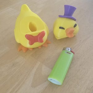 Könnte beinhalten: Ein gelbes entenförmiges Objekt mit einer roten Fliege und ein separates gelbes Entenkopfstück mit einem violetten Zylinder, platziert neben einem hellgrünen Bic-Feuerzeug. Der Entenkörper ist hohl und der Kopf hat geschlossene Augen.
