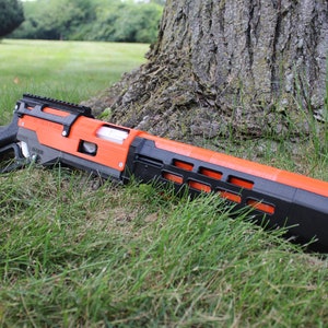 Puede incluir: Un juguete con forma de pistola naranja y negra descansa sobre hierba verde cerca de un tronco de árbol. El juguete tiene un cañón largo con una serie de recortes rectangulares y un mango negro. La pistola es de plástico.