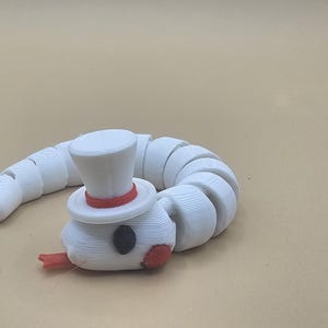 Puede incluir: Una figura de gusano articulado de color blanco con cuerpo segmentado, un pequeño sombrero de copa con una banda roja, un ojo gris oscuro y una lengua roja que sobresale de la boca. El objeto muestra capas de impresión 3D y forma curva.