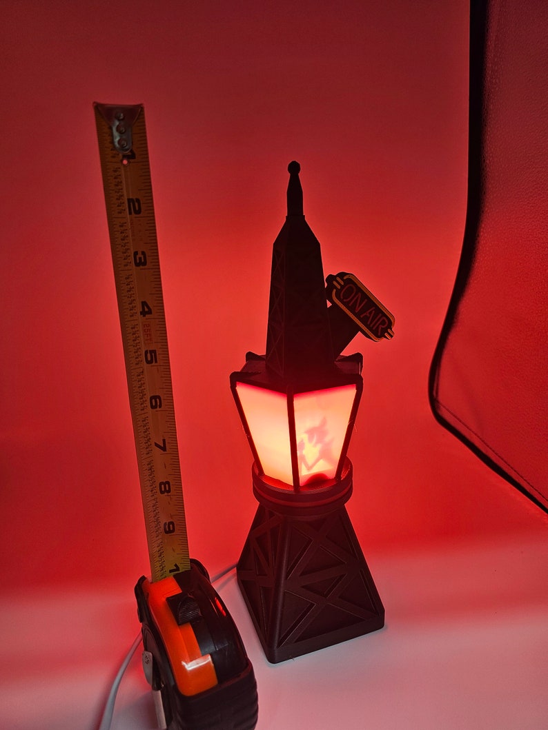 Puede incluir: L&aacute;mpara negra con forma de torre con un brillo rojo, con un letrero de "ON AIR". La l&aacute;mpara mide aproximadamente 18 cm de alto. La base tiene un dise&ntilde;o geom&eacute;trico. Una cinta m&eacute;trica est&aacute; al lado de la l&aacute;mpara.