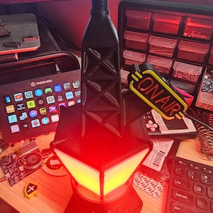 Puede incluir: Una l&aacute;mpara negra y geom&eacute;trica con un resplandor rojo y amarillo. La l&aacute;mpara tiene una estructura similar a una torre y una base en forma de diamante. Las palabras "ON AIR" son visibles en un letrero amarillo. Un Stream Deck y un teclado tambi&eacute;n est&aacute;n en la imagen.