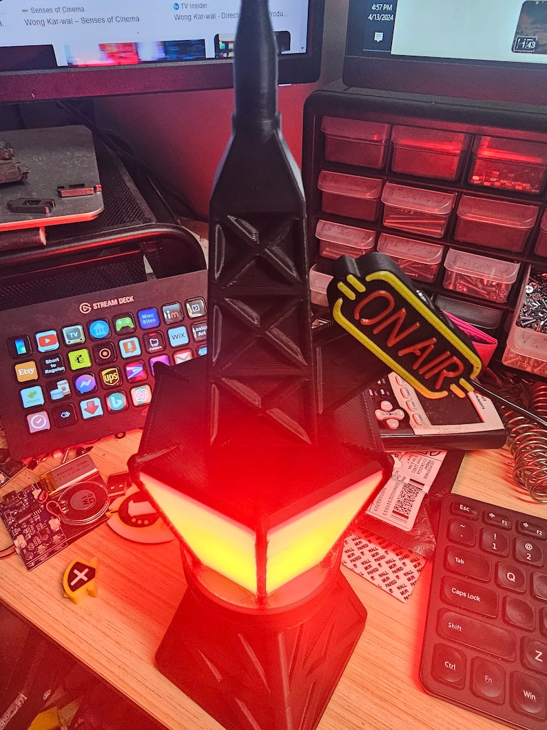 Puede incluir: Una l&aacute;mpara negra y geom&eacute;trica con un resplandor rojo y amarillo. La l&aacute;mpara tiene una estructura similar a una torre y una base en forma de diamante. Las palabras "ON AIR" son visibles en un letrero amarillo. Un Stream Deck y un teclado tambi&eacute;n est&aacute;n en la imagen.