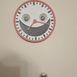 May include: A whimsical wall clock with a smiling face design. The clock has a white face, a coral-colored frame, and coral-colored clock hands. The clock's face features two eye-shaped cutouts and a wide, smiling mouth.