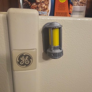 May include: A gray 3D printed GE refrigerator light switch cover with a yellow light inside. The switch cover is attached to a white refrigerator door.